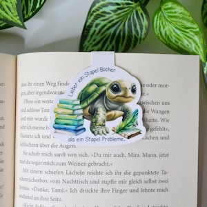 Könnte beinhalten: Ein skurriles Lesezeichen mit einer Zeichentrickschildkröte und einem Stapel Bücher. Die Schildkröte ist grün und braun, mit großen Augen. Das Lesezeichen hat deutschen Text und befindet sich in einem offenen Buch.