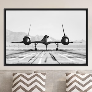 Op de afbeelding: Zwart-wit ingelijste print van een SR-71 Blackbird-vliegtuig op een landingsbaan. Het verkenningsvliegtuig staat centraal, met zijn kenmerkende vorm en lange vleugels zichtbaar. De achtergrond toont bergen en een heldere lucht.