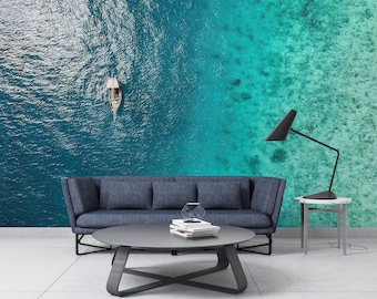 May include: An aerial view of a boat sailing on turquoise water, with a gradient of colors from deep blue to light turquoise. The image is a wall mural behind a blue sofa, a gray coffee table, and a black lamp.