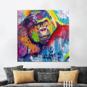 Peut inclure: Une œuvre d'art carrée et vibrante représentant un gorille dans un style graffiti coloré. Le visage du gorille est détaillé avec une expression pensive, et l'œuvre incorpore des teintes vives de rouge, de bleu, de jaune et de violet. L'œuvre est affichée au-dessus d'un canapé gris avec des coussins jaunes.