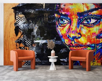 May include: A colorful abstract mural of a woman's face with blue eyes, painted in a graffiti style. Two orange chairs and a small white table with a vase of dried flowers are in front of the mural.
