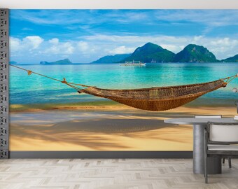 May include: A woven hammock hangs between two trees on a tropical beach. The hammock is made of natural materials and has a rustic look. The beach is sandy and the water is turquoise. Lush green mountains are in the background.