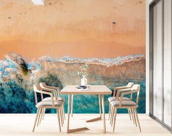 May include: A dining room with a beach-themed mural on the wall. The mural features a sandy beach with turquoise ocean waves crashing on the shore. A wooden table with four chairs is in the foreground.