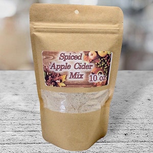 May include: A brown paper stand-up pouch containing Spiced Apple Cider Mix, 10 oz. The label features a wood-grain background with images of apples and autumn leaves. A clear window at the bottom reveals the light-colored, granular powder inside.
