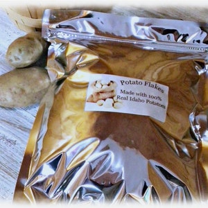 Op de afbeelding: Een zilveren foliezak met aardappelvlokken. De zak heeft een etiket met de tekst "Potato Flakes Made with 100% Real Idaho Potatoes". Op de achtergrond zijn twee hele aardappelen te zien.
