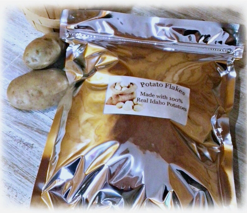 Op de afbeelding: Een zilveren foliezak met aardappelvlokken. De zak heeft een etiket met de tekst "Potato Flakes Made with 100% Real Idaho Potatoes". Op de achtergrond zijn twee hele aardappelen te zien.