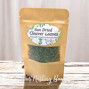 May include: A brown paper bag with a clear window containing dried green cleaver leaves. The bag has an oval label that reads "Sun Dried Cleaver Leaves, Naturally Grown & Pesticide Free, 1/2 c."
