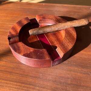 May include: A round, wooden ashtray with a cigar resting on it. The ashtray is made of dark brown wood with a red accent. The cigar is brown and cylindrical. The ashtray is sitting on a wooden surface.