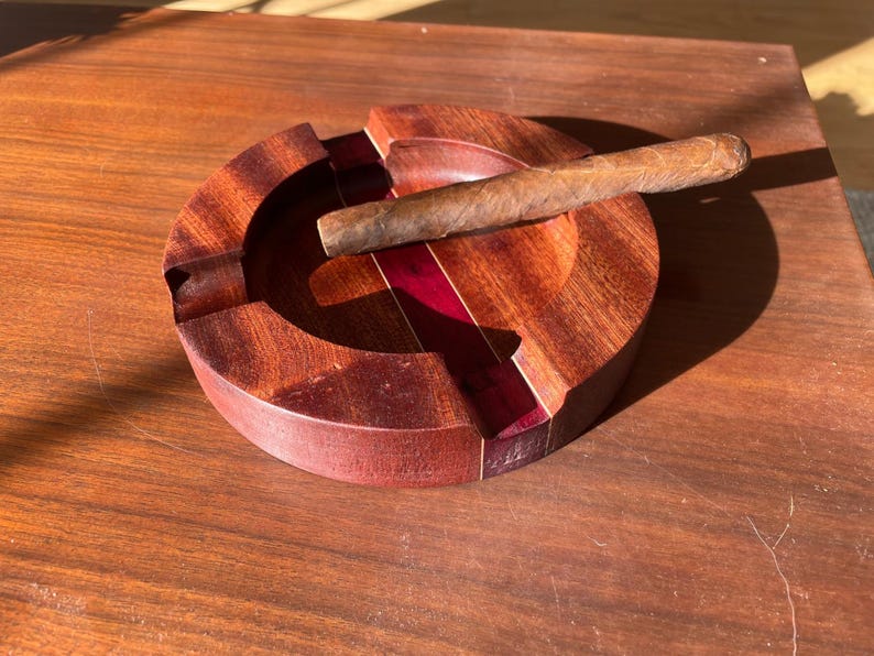 May include: A round, wooden ashtray with a cigar resting on it. The ashtray is made of dark brown wood with a red accent. The cigar is brown and cylindrical. The ashtray is sitting on a wooden surface.