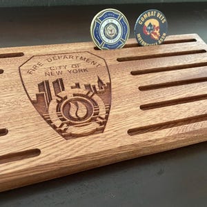 May include: A wooden display stand with slots, featuring the "Fire Department City of New York" emblem and two challenge coins. One coin has a blue and gold design, the other a skull and crossbones. The stand is made of reddish-brown wood.