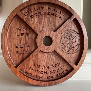 May include: A round, wooden weight plate with engraved text. The plate is brown with a central hole and four cut-out sections. Text includes "45 LBS" and "20.4 KGS", along with names, dates, and a unit logo.