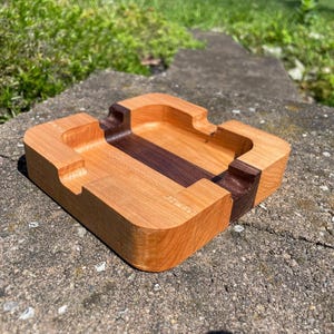 May include: A rectangular wooden ashtray with a light brown exterior and a dark brown center stripe. The ashtray has curved edges and grooves for resting cigarettes. The wood grain is visible.