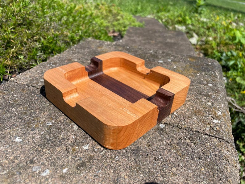 May include: A rectangular wooden ashtray with a light brown exterior and a dark brown center stripe. The ashtray has curved edges and grooves for resting cigarettes. The wood grain is visible.