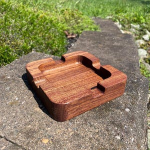 May include: A rectangular wooden ashtray with a rich brown finish. The ashtray features a recessed area for holding ashes and grooves for resting cigarettes. The ashtray is made of solid wood and is designed for outdoor or indoor use.
