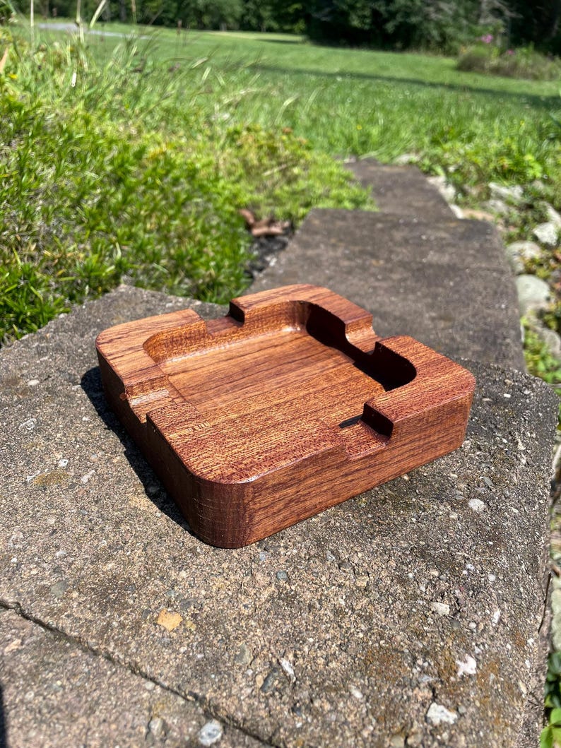 May include: A rectangular wooden ashtray with a rich brown finish. The ashtray features a recessed area for holding ashes and grooves for resting cigarettes. The ashtray is made of solid wood and is designed for outdoor or indoor use.