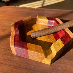 May include: A wooden ashtray with a cigar resting on it. The ashtray is a geometric shape with alternating stripes of red, yellow, and brown wood. The cigar is brown and cylindrical. The ashtray is on a wooden surface.
