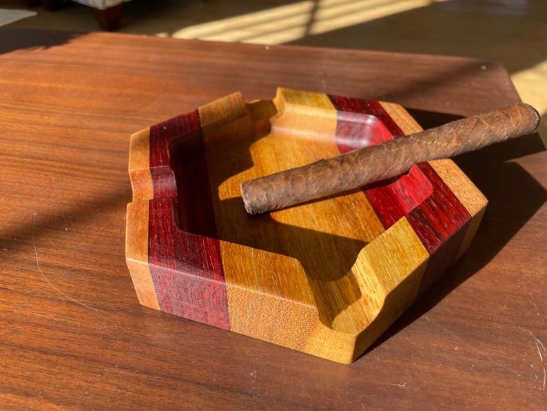 May include: A wooden ashtray with a cigar resting on it. The ashtray is a geometric shape with alternating stripes of red, yellow, and brown wood. The cigar is brown and cylindrical. The ashtray is on a wooden surface.