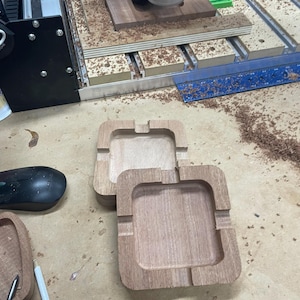 May include: Two unfinished wooden ashtrays with square shapes and rounded corners sit on a light brown surface. The ashtrays have a recessed area for holding ashes and a groove for resting a cigarette. Wood shavings are scattered around the ashtrays.