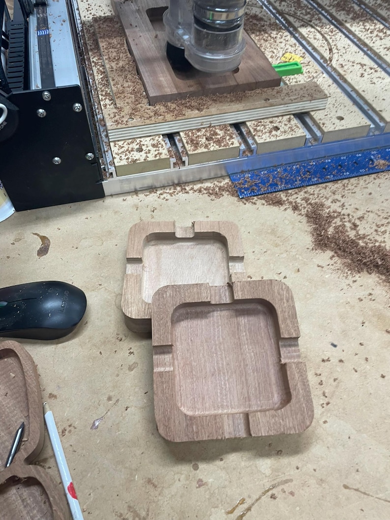 May include: Two unfinished wooden ashtrays with square shapes and rounded corners sit on a light brown surface. The ashtrays have a recessed area for holding ashes and a groove for resting a cigarette. Wood shavings are scattered around the ashtrays.