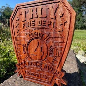 May include: A wooden plaque with a red-brown finish, depicting a fire department badge. The badge features the text "TROY FIRE DEPT." and "ENGINE 24" with a brick pattern background. The text "THE BRICK OVEN Charles Eddy" is displayed on a banner below the badge.