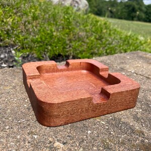 May include: A square, wooden ashtray with a rich, reddish-brown finish. The ashtray has a recessed center and four curved rests for cigarettes. The ashtray is made of solid wood and has a smooth, polished surface.