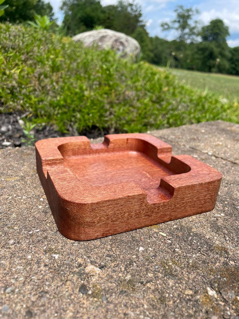 May include: A square, wooden ashtray with a rich, reddish-brown finish. The ashtray has a recessed center and four curved rests for cigarettes. The ashtray is made of solid wood and has a smooth, polished surface.
