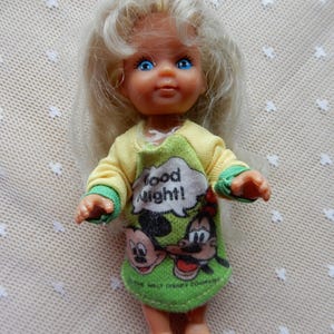 Könnte beinhalten: Eine kleine blonde Puppe, die ein grünes und gelbes Nachthemd mit Mickey Mouse und Goofy trägt. Das Nachthemd trägt den Schriftzug "Good Night!"