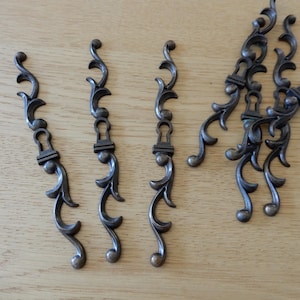 Könnte beinhalten: Eine Sammlung antiker, bronzefarbener Schrankgriffe mit einem verzierten, geschwungenen Design. Jeder Griff verfügt über ein Schlüssellochdetail und eine dekorative Verzierung an der Basis. Die Hardware ist auf einer hellen Holzoberfläche angeordnet.