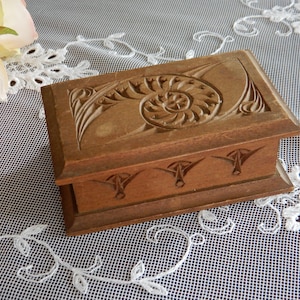 Könnte beinhalten: Eine rechteckige Holzbox mit einem geschnitzten Spiralmuster auf dem Deckel und dekorativen Schnitzereien auf der Vorderseite. Die Box hat eine warme braune Farbe und liegt auf einer weißen Spitzentischdecke.
