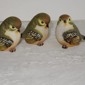 Könnte beinhalten: Drei kleine Keramik-Vogelfiguren. Die Vögel sind in Grün-, Braun- und Weißtönen bemalt. Sie haben realistische Details, darunter Federn und Schnäbel.