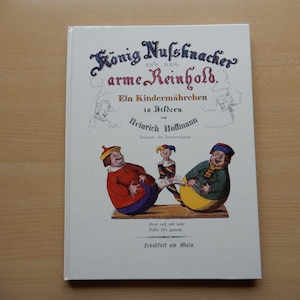Könnte beinhalten: Ein Vintage-Buchcover mit einer farbenfrohen Illustration von drei Figuren. Der Titel lautet "König Nussknacker und der arme Reinhold." Das Buch ist ein Kindermärchen in Bildern von Heinrich Hoffmann.