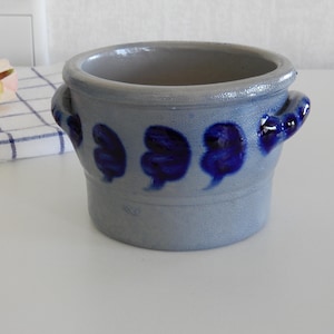 Könnte beinhalten: Kleiner, grauer Keramiktopf mit zwei Henkeln und dunkelblauen Blumenmustern. Der Topf hat eine runde Form und steht auf einer weißen Oberfläche. Eine Rose und ein kariertes Tuch befinden sich im Hintergrund.