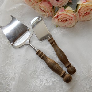 Könnte beinhalten: Zwei Käsemesser mit Holzgriffen. Die Messer liegen auf einer weißen Spitzentischdecke. Ein Messer ist eine Reibe und das andere ist ein Buttermesser.