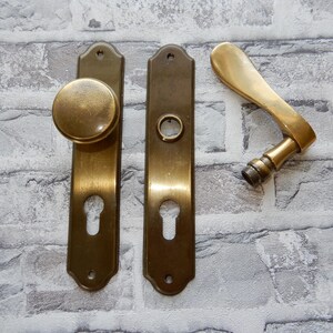 Könnte beinhalten: Antikes Türbeschlagsset aus Messing, bestehend aus einem Türknauf, einem Griff und zwei Rosetten. Die Beschläge haben eine warme, gealterte Patina, mit Schlüssellochausschnitten und einem klassischen Design.