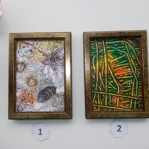 Könnte beinhalten: Zwei kleine gerahmte Kunstwerke mit goldfarbenen Rahmen. Eines zeigt ein farbenfrohes abstraktes Design mit floralen und geometrischen Elementen. Das andere hat ein grünes, gelbes und oranges abstraktes Design. Beide befinden sich auf einer weißen Oberfläche.