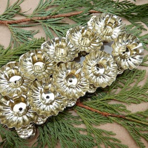 Könnte beinhalten: Eine Ansammlung von vintage, goldfarbenen Kerzenhaltern mit einem gerüschten Randdesign. Die Kerzenhalter sind auf einem Bett aus grünen Tannenzweigen angeordnet und schaffen eine festliche und dekorative Präsentation. Die Kerzenhalter sind aus Metall.