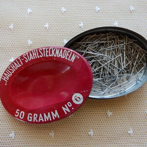 Könnte beinhalten: Eine rote Metalldose mit einem weißen Etikett, auf dem "Haushalt-Stahlstecknadeln" 50 Gramm Nr. 6 steht. Die Dose ist geöffnet und mit silbernen Stecknadeln gefüllt.