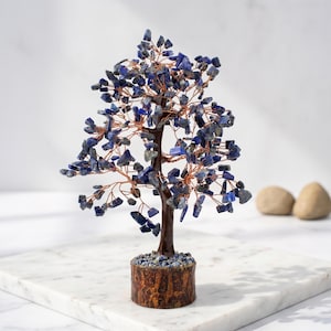 May include: A decorative tree sculpture featuring numerous small, tumbled blue lapis lazuli stones attached to thin, twisted copper wire branches. The tree has a dark brown, textured trunk set into a cylindrical base covered in small blue stones.