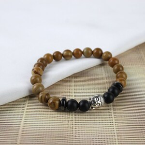 Puede incluir: Un brazalete de cuentas marrones y negras con un dije de cabeza de Buda plateado. El brazalete está hecho de cuentas de piedra natural y tiene un separador de cuentas negro.
