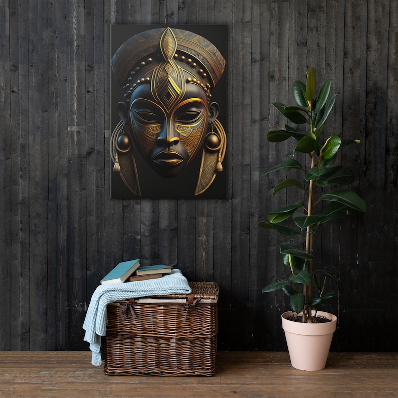 May include: A canvas print featuring a detailed, stylized African mask. The mask is primarily black with intricate gold-colored accents and geometric patterns. The artwork is displayed on a dark wooden wall, with a wicker basket and a potted plant in the foreground.