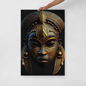 May include: A black canvas print featuring a detailed, stylized African mask. The mask has intricate gold-colored patterns and accents, including a headdress, facial details, and dangling earrings. The artwork is held up by a person's hand.