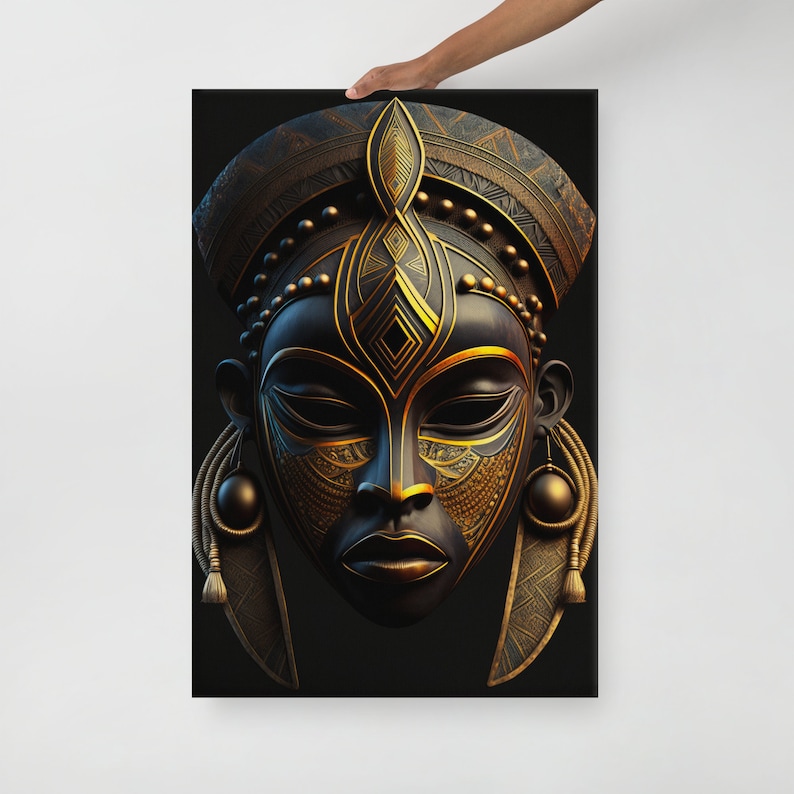 May include: A black canvas print featuring a detailed, stylized African mask. The mask has intricate gold-colored patterns and accents, including a headdress, facial details, and dangling earrings. The artwork is held up by a person's hand.