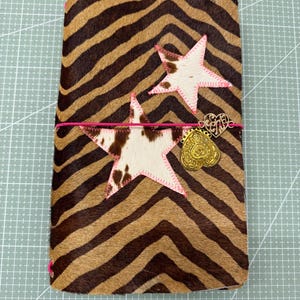 Könnte beinhalten: Ein braun-beiger Zebrastreifen-Journal-Einband mit zwei sternförmigen Aufnähern. Die Sterne sind weiß mit braunen Flecken und rosa Nähten. Ein rosa Gummiband und ein goldfarbener Anhänger mit Herzdesign vervollständigen den Einband.