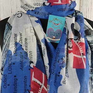 May include: A white scarf featuring a printed map of England. The map is in blue, red and white. The scarf is folded and draped over a wooden surface. The text on the scarf includes the names of cities and towns in England, such as London, Bristol and Southampton.
