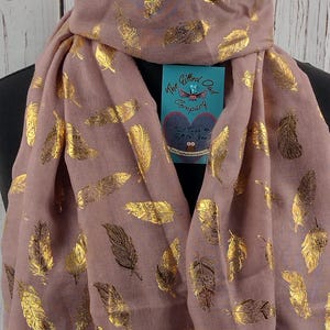 May include: A blush pink scarf with a gold foil feather pattern. The scarf has a tag attached that reads "The Gifted Owl Company" and "Treat Yourself to a Gift for You".