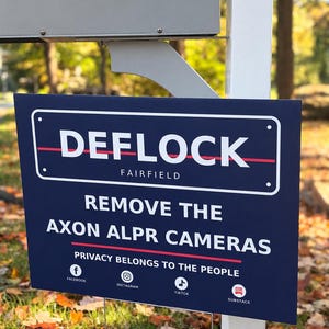 May include: A rectangular dark blue yard sign with white text reading DEFLOCK FAIRFIELD, REMOVE THE AXON ALPR CAMERAS, and PRIVACY BELONGS TO THE PEOPLE. The sign features a thin red horizontal line and icons for Facebook, Instagram, TikTok, and Substack.