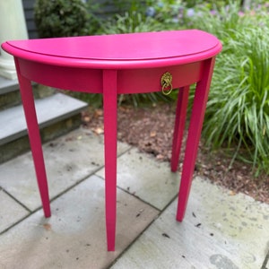 May include: A pink half-moon table with a single drawer and a gold lion head pull. The table has three legs and is sitting on a gray stone patio.