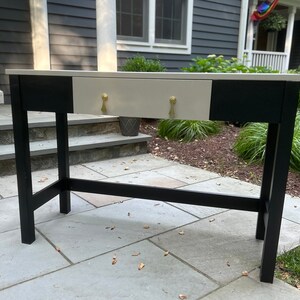 May include: A black and white console table with a single drawer. The drawer has gold-colored pulls. The table is standing on a stone patio.