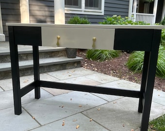 May include: A black and white console table with a single drawer. The drawer has gold-colored pulls. The table is standing on a stone patio.