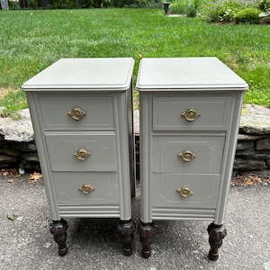 May include: Two matching gray painted nightstands with three drawers each. The drawers have gold-toned hardware. The nightstands have dark wood legs.